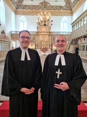 Zwei Männer im schwarzen Talar, einer hat ein Kreuz umgehängt