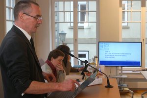 Ein Mann steht an einem Rednerpult mit Mikro, im Hintergrund sitzen Menschen an einem Tisch und ein Bildschirm ist sichtbar