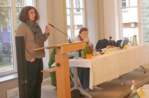 Eine Frau steht an einem Rednerpult, daneben sitzt eine Frau an einem Tisch