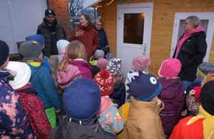 Viele winterlich gekleidete Kinder sitzen mit dem Rücken zur Kamera, vier Erwachsene stehen den Kindern zugewandt vorne