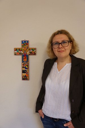 Eine junge Frau mit lockigen Haaren und einer Brille steht neben einem bunten Kreuz, welches an der Wand hängt