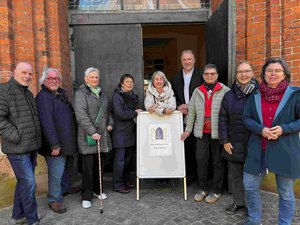 Eine Gruppe von Menschen steht mit einem Aufsteller vor einer offenen Kirchentür.